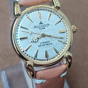 May include: A gold-toned Jaeger-LeCoultre Club watch with a light-colored face and a brown leather strap. The watch face features gold-colored hands and markers, with the text "17 JEWELS SHOCK PROOF". The watch has a round face and a light brown leather strap.