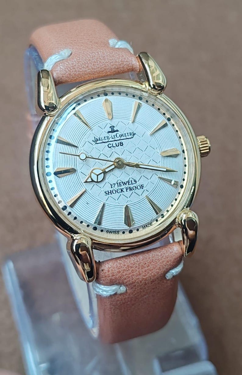 May include: A gold-toned Jaeger-LeCoultre Club watch with a light-colored face and a brown leather strap. The watch face features gold-colored hands and markers, with the text "17 JEWELS SHOCK PROOF". The watch has a round face and a light brown leather strap.