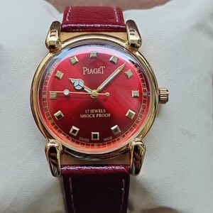 May include: A gold-toned Piaget wristwatch with a vibrant red dial and a matching leather band. The watch face features gold markers and hands, with the brand name prominently displayed. The watch is marked "17 Jewels Shock Proof" and "Swiss Made".