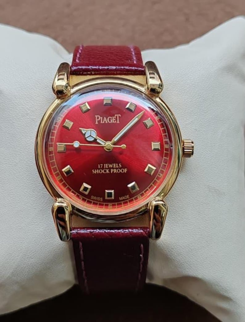 May include: A gold-toned Piaget wristwatch with a vibrant red dial and a matching leather band. The watch face features gold markers and hands, with the brand name prominently displayed. The watch is marked "17 Jewels Shock Proof" and "Swiss Made".