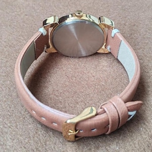 May include: A gold-toned watch with a light brown leather band. The watch face is not visible. The band is secured with a gold-colored buckle. The watch is set against a neutral background.
