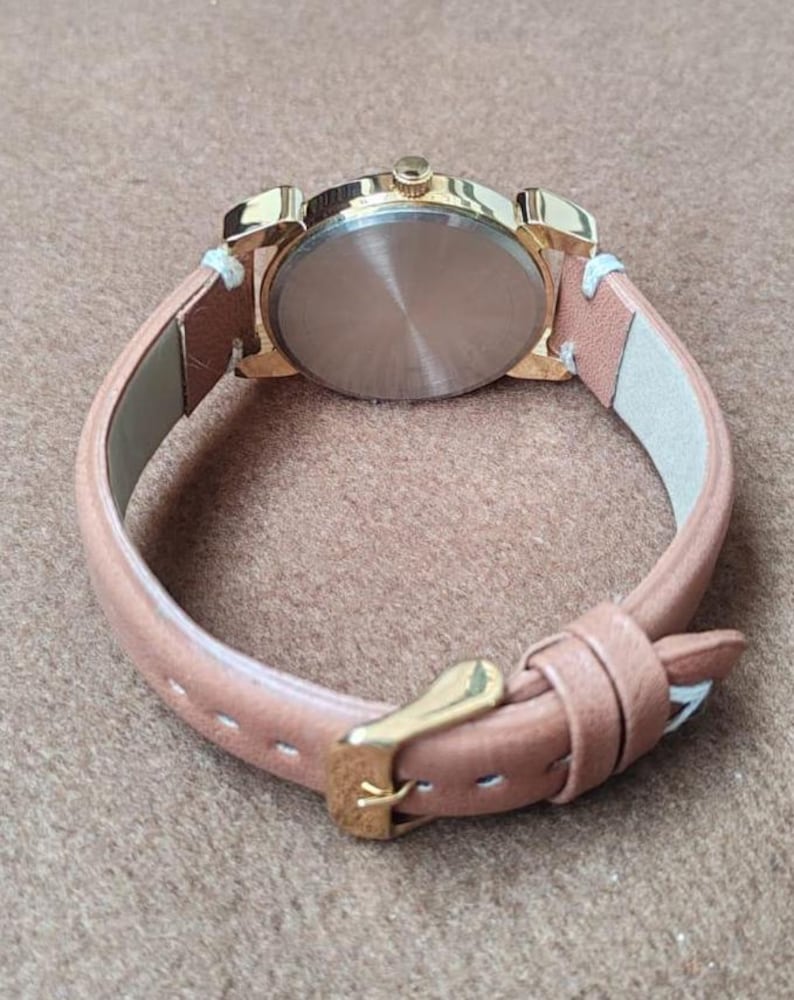 May include: A gold-toned watch with a light brown leather band. The watch face is not visible. The band is secured with a gold-colored buckle. The watch is set against a neutral background.