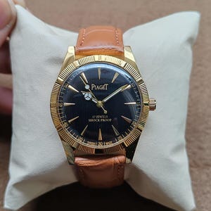 May include: A gold-toned Piaget wristwatch with a black face and a brown leather band. The watch face displays the brand name and the text "17 Jewels Shock Proof". The watch is displayed on a cream-colored pillow.