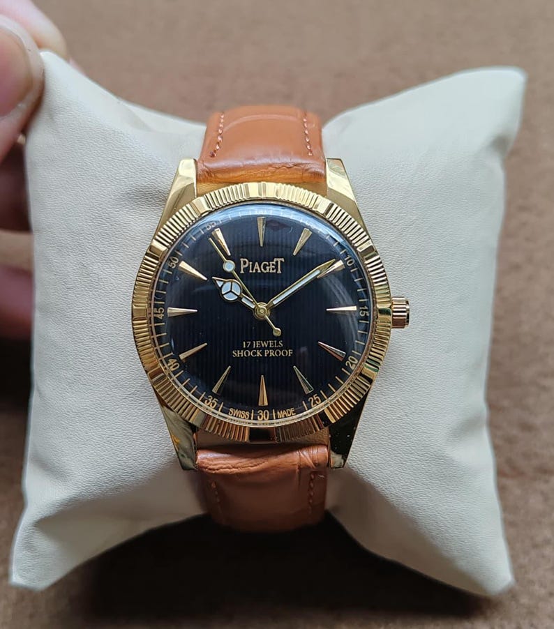 May include: A gold-toned Piaget wristwatch with a black face and a brown leather band. The watch face displays the brand name and the text "17 Jewels Shock Proof". The watch is displayed on a cream-colored pillow.