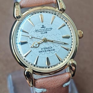 May include: A gold-toned Jaeger-LeCoultre Club watch with a light-colored face and a brown leather strap. The watch face features the brand name, the word "CLUB", and the text "17 JEWELS SHOCK PROOF".
