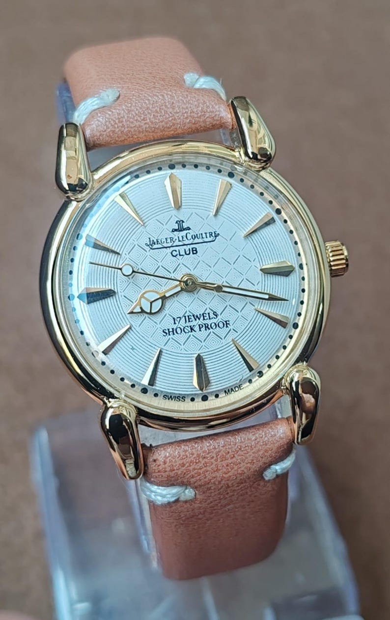 May include: A gold-toned Jaeger-LeCoultre Club watch with a light-colored face and a brown leather strap. The watch face features the brand name, the word "CLUB", and the text "17 JEWELS SHOCK PROOF".