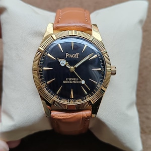 May include: A gold-toned Piaget wristwatch with a black face and gold hands. The watch has a brown leather band and is marked "17 JEWELS SHOCK PROOF". The watch is resting on a white pillow.