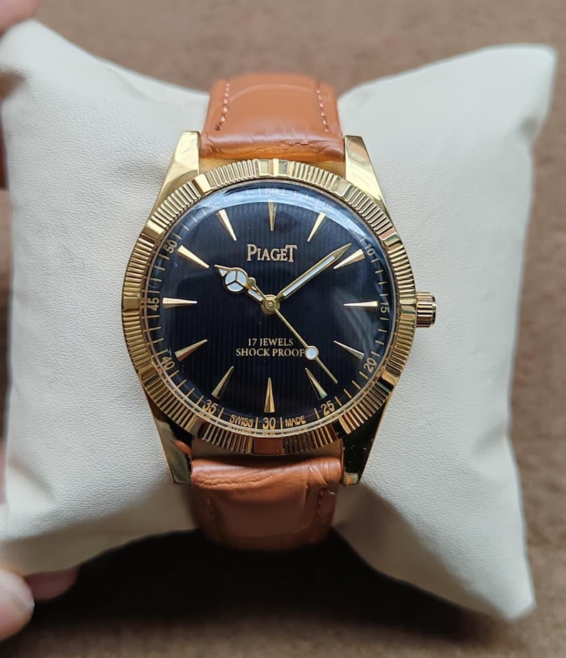May include: A gold-toned Piaget wristwatch with a black face and gold hands. The watch has a brown leather band and is marked "17 JEWELS SHOCK PROOF". The watch is resting on a white pillow.