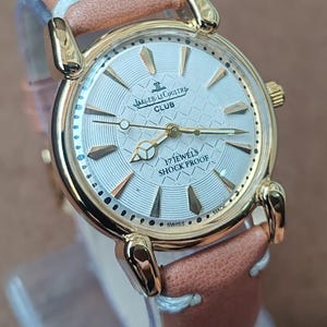 May include: A gold-toned Jaeger-LeCoultre Club watch with a light-colored face and a brown leather strap. The watch face features gold-colored hands and markers, with the text "17 JEWELS SHOCK PROOF" and "SWISS MADE".
