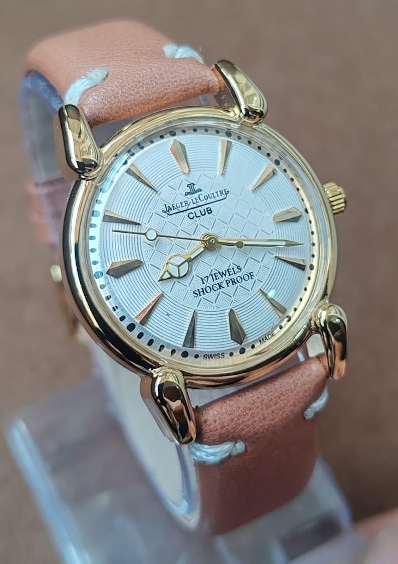May include: A gold-toned Jaeger-LeCoultre Club watch with a light-colored face and a brown leather strap. The watch face features gold-colored hands and markers, with the text "17 JEWELS SHOCK PROOF" and "SWISS MADE".