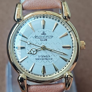 May include: A gold-toned Jaeger-LeCoultre Club watch with a light brown leather strap. The watch face is silver with gold markers and hands. The text "17 JEWELS SHOCK PROOF" is visible on the watch face. The watch is Swiss made.
