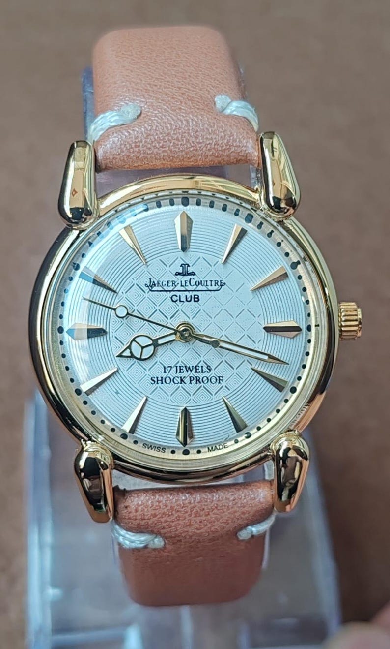 May include: A gold-toned Jaeger-LeCoultre Club watch with a light brown leather strap. The watch face is silver with gold markers and hands. The text "17 JEWELS SHOCK PROOF" is visible on the watch face. The watch is Swiss made.