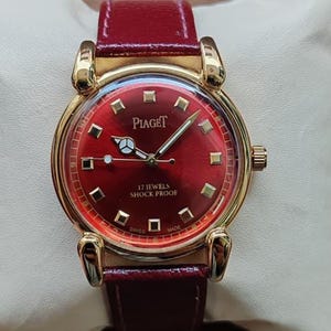 May include: A gold-toned Piaget wristwatch with a deep red face and matching leather band. The watch face features gold markers and hands, with the text "17 Jewels Shock Proof" and "Swiss Made". The watch is displayed on a cream-colored surface.