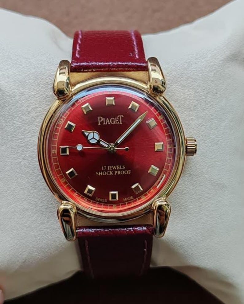 May include: A gold-toned Piaget wristwatch with a deep red face and matching leather band. The watch face features gold markers and hands, with the text "17 Jewels Shock Proof" and "Swiss Made". The watch is displayed on a cream-colored surface.