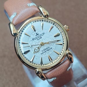 May include: A gold-toned Jaeger-LeCoultre Club watch with a light-colored face and brown leather band. The watch face features the text "17 JEWELS SHOCK PROOF" and "SWISS MADE". The watch has gold-colored hands and hour markers.