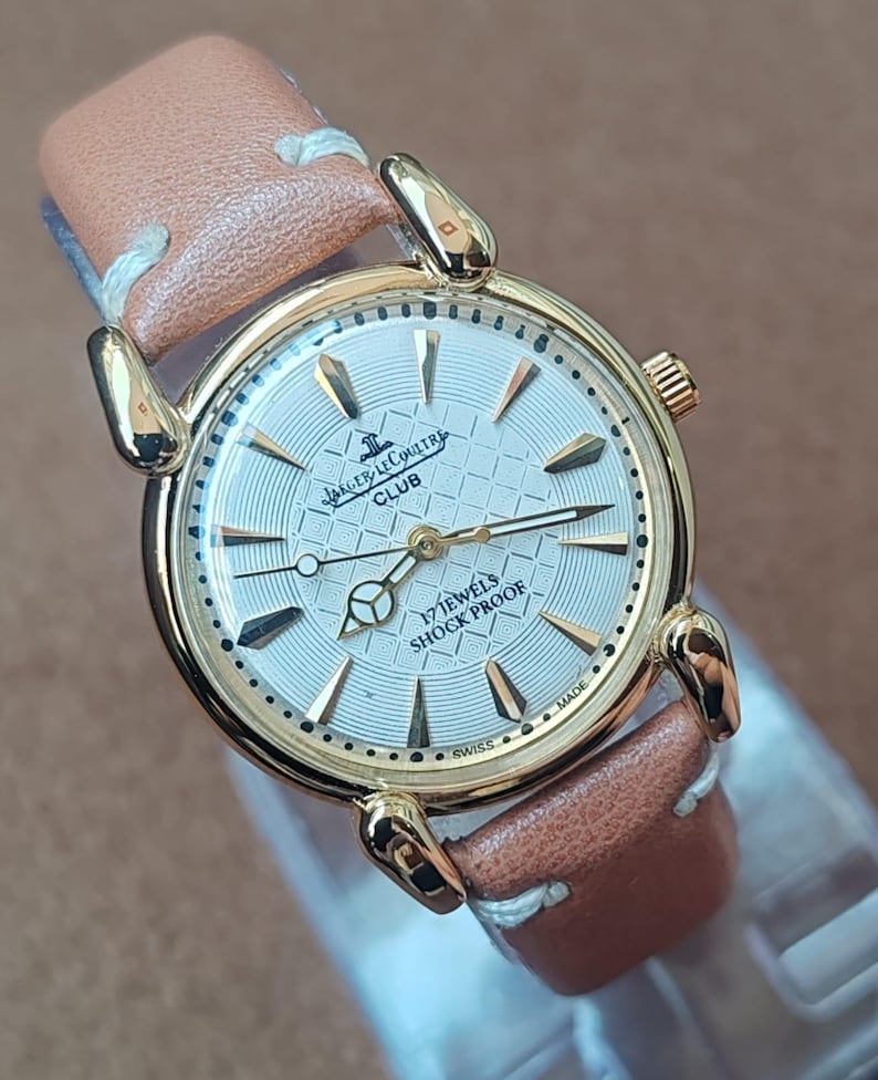 May include: A gold-toned Jaeger-LeCoultre Club watch with a light-colored face and brown leather band. The watch face features the text "17 JEWELS SHOCK PROOF" and "SWISS MADE". The watch has gold-colored hands and hour markers.