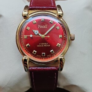 May include: A gold-toned Piaget wristwatch with a vibrant red face and a matching burgundy leather band. The watch features gold hands and markers, and the face displays the brand name and "17 JEWELS SHOCK PROOF".
