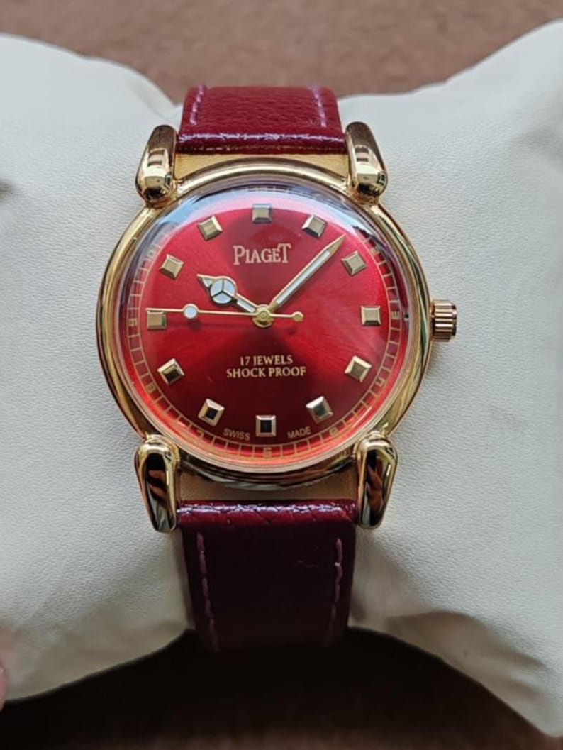 May include: A gold-toned Piaget wristwatch with a vibrant red face and a matching burgundy leather band. The watch features gold hands and markers, and the face displays the brand name and "17 JEWELS SHOCK PROOF".