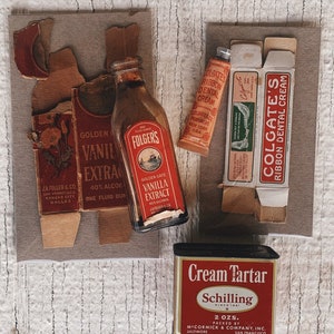 May include: A collection of vintage kitchen and bathroom items, including a bottle of Folgers Vanilla Extract, a tube of Colgate Ribbon Dental Cream, and a tin of Schilling Cream Tartar. The items are arranged on a light brown wooden surface.