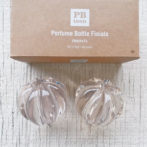 Puede incluir: Un juego de dos embellecedores para botellas de perfume de vidrio transparente. Los embellecedores tienen forma de flor con una textura acanalada. Los embellecedores están empaquetados en una caja marrón con el texto "PB teen Perfume Bottle Finials Embouts Set of two / une paire" impreso en la caja.