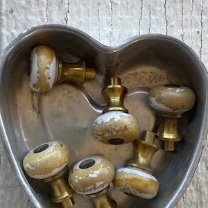 Könnte beinhalten: Eine Sammlung von sieben antiken Keramik-Schrankknöpfen mit Messingbeschlägen. Die Knöpfe sind weiß mit einer abgenutzten goldenen Oberfläche und haben eine runde Form. Die Knöpfe befinden sich in einem herzförmigen Metallbehälter.