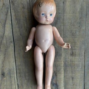 Puede incluir: Una muñeca vintage con rostro pintado, ojos azules y cabello castaño claro. La muñeca tiene un cuerpo desnudo y está posada sobre una superficie de madera envejecida. Las extremidades de la muñeca están ligeramente articuladas y la pintura muestra signos de edad.