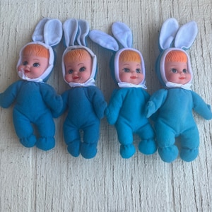 Puede incluir: Cuatro muñecas de peluche de conejo azul con orejas blancas y ribetes blancos. Cada muñeca tiene una cara sonriente con cabello naranja y ojos azules.