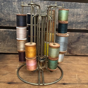 Puede incluir: Un antiguo soporte metálico para carretes de hilo lleno de carretes de hilo de varios colores. Los carretes son en tonos de negro, marrón, dorado, rosa, verde y gris. El soporte tiene una base circular y múltiples brazos para sujetar los carretes.