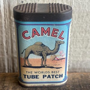 May include: Vintage Camel tube patch tin. The tin features a blue background with the word "CAMEL" in red lettering. A camel is depicted on the front, with the text "THE WORLDS BEST TUBE PATCH" below. The tin has a black top.