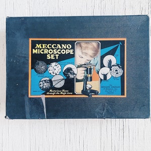 Puede incluir: Una caja vintage azul y negra para un juego de microscopio Meccano. La caja presenta a un niño mirando a través del microscopio e imágenes de objetos magnificados como el ojo de una mosca, la pata de una araña y una escama de ala de polilla. El texto en la caja dice "Meccano Microscope Set" y "Mysterious Views through the Magic Lens".