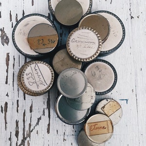 Może przedstawiać: Kolekcja vintage metalowych puszek z różnymi etykietami i oznaczeniami. Puszki są okrągłe i mają srebrno-złoto-biały schemat kolorystyczny. Niektóre z etykiet mają ręcznie napisany tekst, w tym "8.5W" i "Eterna".