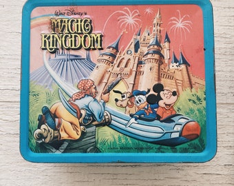 Op de afbeelding: Vintage Walt Disney's Magic Kingdom lunchbox met een blauwe achtergrond en een kleurrijke illustratie van Assepoester's kasteel, Space Mountain en Mickey Mouse, Donald Duck en Captain Jack Sparrow.