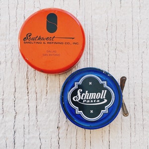May include: Two vintage metal tins. The first tin is red with black text that reads "Southwest Smelting & Refining Co., Inc. Dallas San Antonio". The second tin is blue with a white label that reads "Schmoll Pasta" in a cursive font.