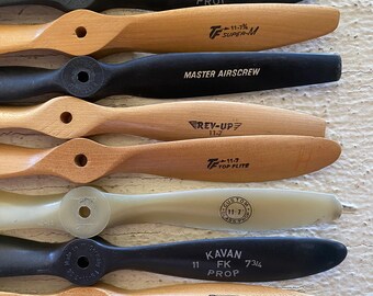 May include: A collection of eight wooden and black airplane propellers. The propellers are various sizes and have different brand names and model numbers printed on them, including "Kavan", "TF Super-M", "Rev-Up", "Top Flite", "Custom Class-A Prop", and "Zinger Pusher".