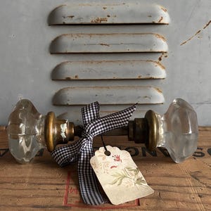 Puede incluir: Dos pomos de puerta antiguos de cristal con herrajes de latón están atados con una cinta a cuadros en blanco y negro. Una etiqueta floral está adjunta. Los pomos de las puertas se exhiben sobre un fondo de metal desgastado y una superficie de madera.