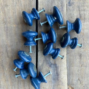 Puede incluir: Una colección de diez pomos de armario azules con tornillos plateados. Los pomos están hechos de madera y tienen un acabado desgastado.