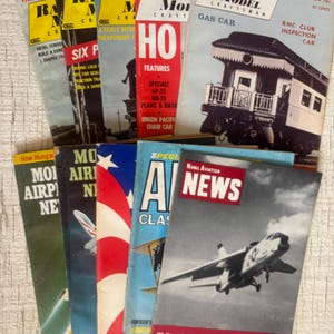 Op de afbeelding: Een verzameling vintage tijdschriften met titels als "Railroad Model Craftsman" en "Naval Aviation News." De covers tonen illustraties van treinen en vliegtuigen. De tijdschriften zijn op elkaar gestapeld, met een verscheidenheid aan vintage publicaties.