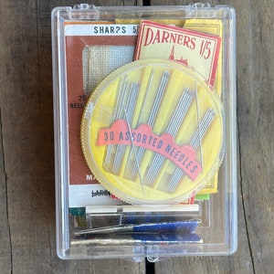 May include: A clear plastic box containing a variety of sewing needles. The box includes a round container with 30 assorted needles, a small piece of fabric, and two cards with the text "SHARP'S 5" and "DARNERS 1/5".