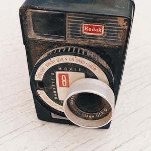 May include: A vintage black Kodak Hawkeye 8 movie camera. The camera has a silver lens and a dial with settings for different lighting conditions. The Kodak logo is visible on the front.