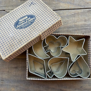 May include: A vintage box of metal cake and sandwich cutters in the shape of playing card suits and a star. The box is brown and white gingham and has the text "Fagley Cake and Sandwich Cutters for Ladies' Card Party Teas, Luncheons, Etc."