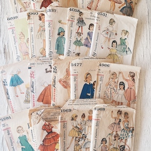 May include: A collection of vintage sewing patterns for children's clothing. The patterns are printed on paper and feature illustrations of girls wearing dresses, skirts, and other garments. The patterns are from the 1960s and 1970s and are in various colours, including pink, blue, green, and yellow.