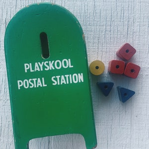 May include: Green wooden Playskool Postal Station toy with a slot in the top.  The toy is accompanied by a set of colorful wooden blocks, including red, yellow, and blue.