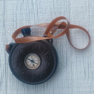 May include: A brown leather canteen with a compass on the front and a brown leather strap.