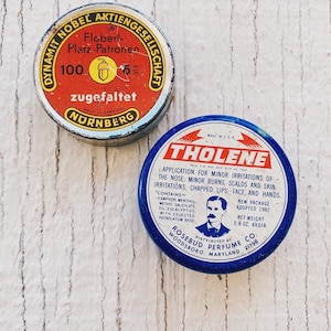May include: Two round metal tins. The tin on the left is red and yellow with German text that reads "Dynamit Nobel Aktiengesellschaft Flobert-Platz-Patronen 100 zugefaltet Nürnberg". The tin on the right is blue and white with the text "Tholene" in red. The text below reads "Application for minor irritations of the nose; minor burns, scalds and skin irritations; chapped lips, face, and hands." The text below that reads "Contains - Camphor, Menthol, Methyl Salicylate, Oil Eucalyptus, with selected petrolatum base." The text below that reads "Distributed by Rosebud Perfume Co. Woodsboro, Maryland 21798".