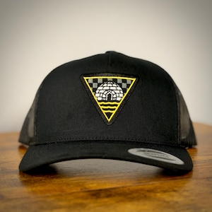 May include: Black trucker hat with a mesh back and a curved brim. The front features a triangular patch with a yellow border, a checkered top, and an igloo design. The hat is on a wooden surface.
