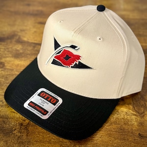 May include: A beige and black baseball cap with a black brim. The cap features a red and black logo on the front and a small black button on top. The label on the brim reads "OTTO Classic Fit".