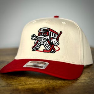 May include: Cream-colored baseball cap with a red brim and a hockey-themed embroidered patch. The patch features a stylized hockey goalie in red, black, and white. The cap has a curved brim and a button on top.