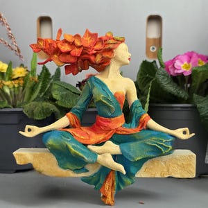 Könnte beinhalten: Skulptur einer Frau im Lotussitz, gekleidet in orange- und türkisfarbene Kleidung und mit einem floralen Kopfschmuck. Die Figur sitzt auf einer beigen Plattform, die Meditation suggeriert. Ein dekoratives Objekt für Haus oder Garten.