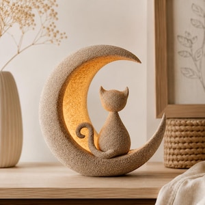 Könnte beinhalten: Eine mondsichelförmige Lampe mit einer texturierten, sandbeigen Oberfläche. Eine kleine Katzenfigur mit passender texturierter Oberfläche sitzt in der Mitte des Mondes. Die innere Kurve des Mondes strahlt ein warmes, sanftes Licht aus.
