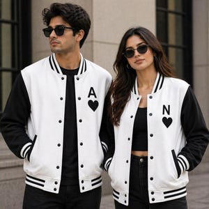 May include: Two varsity jackets featuring a white body with black sleeves and black striped ribbed trim at the collar, cuffs, and hem. The left chest of each jacket displays a black heart icon with a letter, one with an A and one with an N.